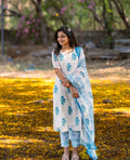 Woman in a white and blue traditional outfit standing in a natural setting with trees and yellow flowers on the ground.